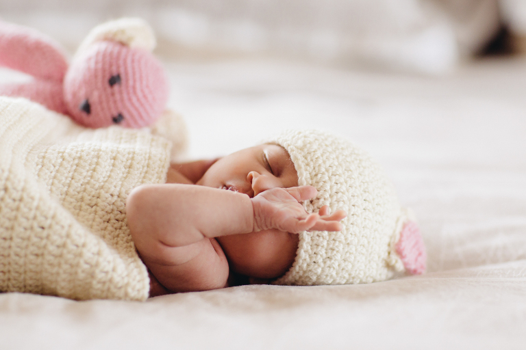 baby girl on bed wrapped in blanket with hat on rubbing face next to a pink stuffed bear Newborn Photography Wellington natural light