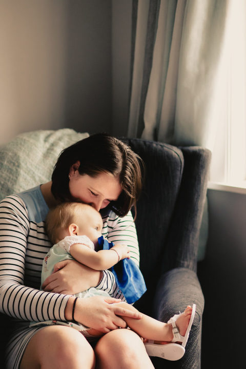 woman kissing baby girl on head staring out the window natural light In-Home Photography First Birthday Party