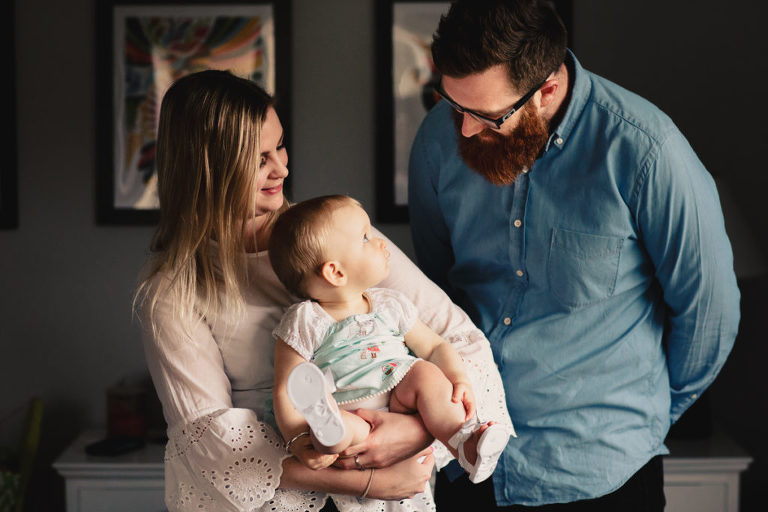 mum holding baby girl while dad looks on natural light In-Home Photography First Birthday Party